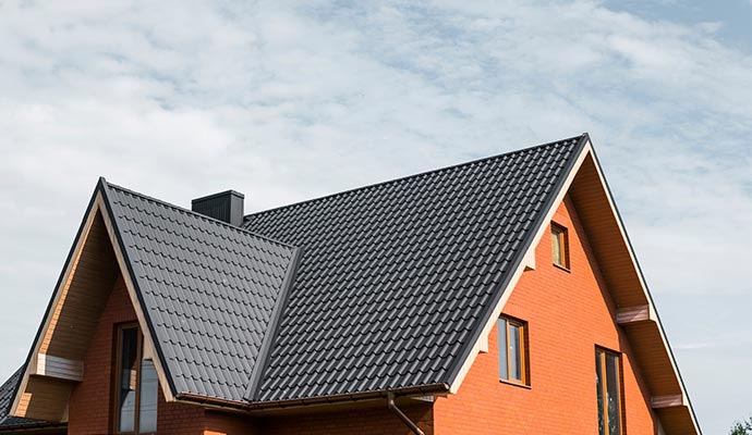A view of a brick house with multiple gables featuring a newly installed, dark grey metal shingle roof under a blue sky