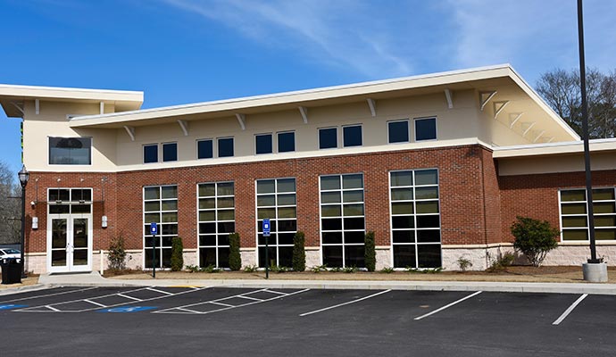A commercial building with brick and beige siding, large windows, and a paved parking lot with marked spaces