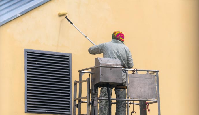 A painter on a vertical platform lift uses a roller on an extension pole to paint the yellow exterior wall of a commercial building.