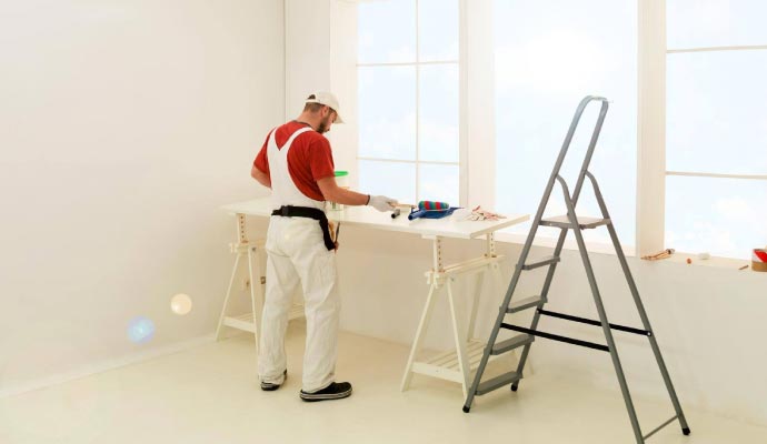 A painter in white overalls stands at a temporary work table, preparing his tools in a bright, white room with a large window.