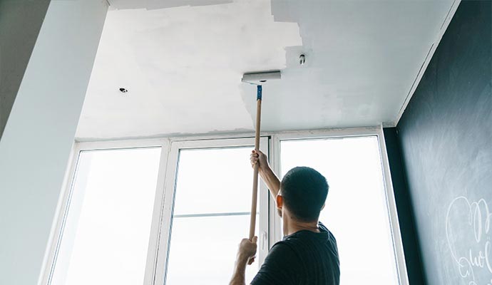A person seen from behind using a paint roller on an extension pole to paint the ceiling of a room white.