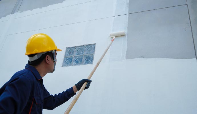 A painter wearing a yellow hard hat uses a roller on an extension pole to apply white primer to the large grey exterior wall of a commercial building.