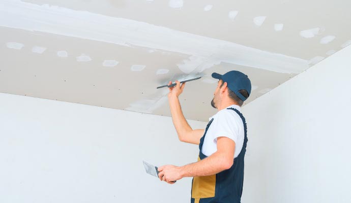 A worker in overalls and a cap uses a taping knife to apply joint compound to the seams of a drywall ceiling.