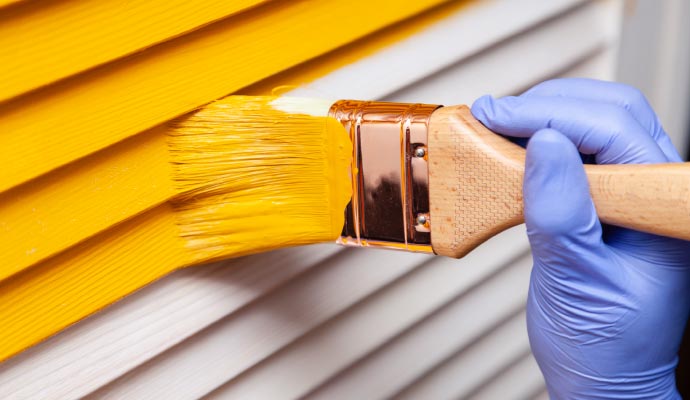 A close-up of a gloved hand using a paintbrush to apply bright yellow paint to the slats of a white louvered door.